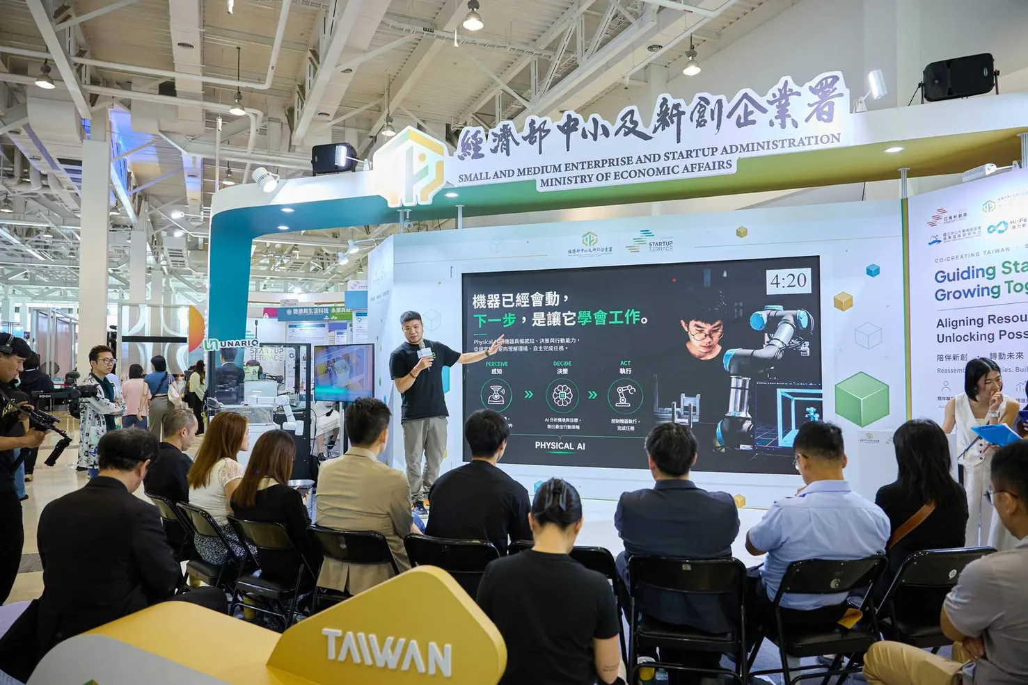 優那科創共同創辦人孫偉強於經濟部中小及新創企業署主題館舞台演講,投影幕標題為「機器已經會動,下一步,是讓它學會工作」,並列出 Physical AI 的感知、決策、執行三個環節